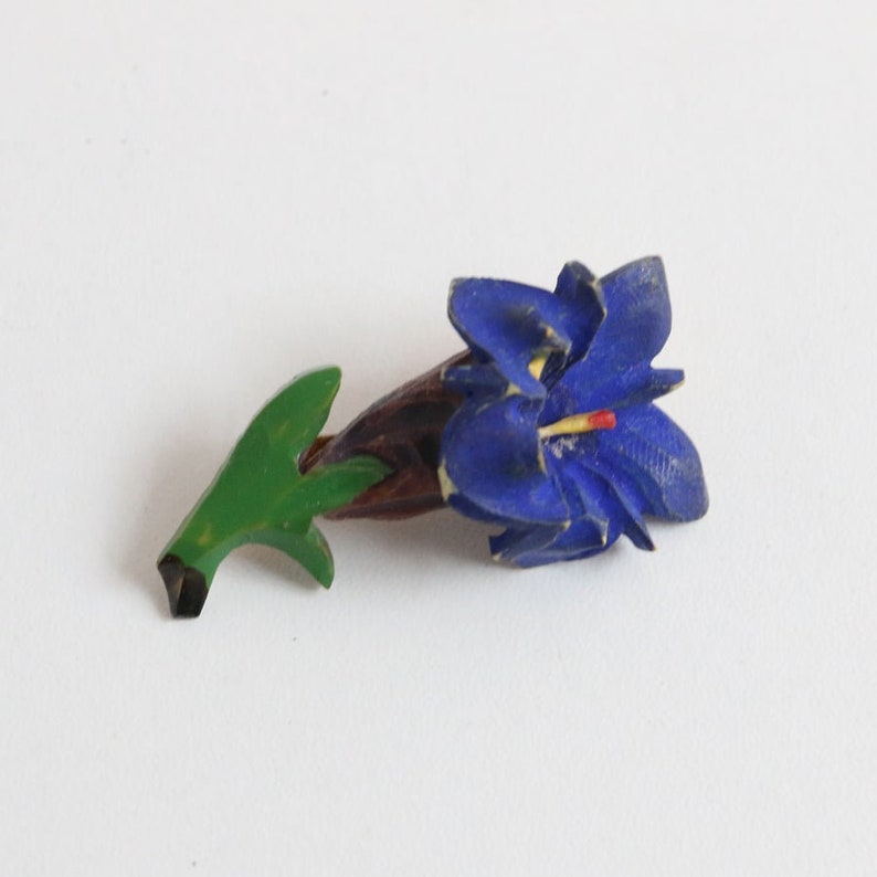 May include: A hand-painted wooden flower brooch. The brooch features a blue flower with yellow and red accents, a green stem, and a brown base. The brooch is a floral accessory.