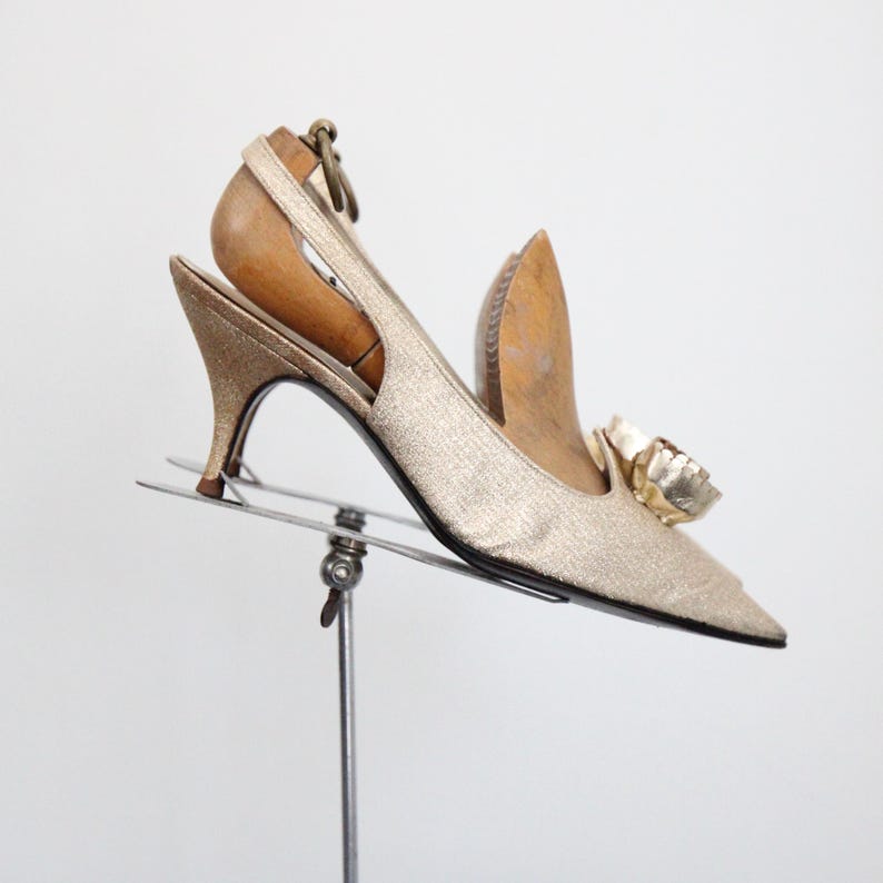 May include: A vintage gold slingback heel with a pointed toe and a small heel. The shoe features a decorative gold flower on the toe and is displayed on a metal stand. The heel height is approximately 2.5 inches.