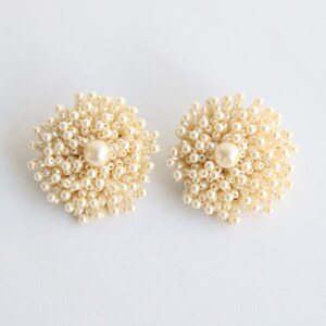 May include: A pair of cream-colored, clustered pearl earrings. Each earring features a large, central pearl surrounded by smaller pearls, creating a floral or starburst design. The earrings are set against a white background.