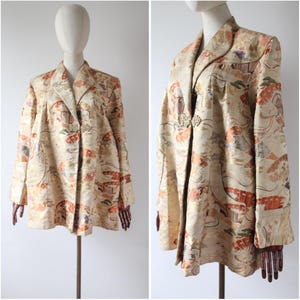 Può includere: Una giacca color crema con un motivo floreale e dettagli dorati. La giacca ha un colletto alla coreana e una chiusura con bottone. Il tessuto è un broccato con una trama in rilievo.