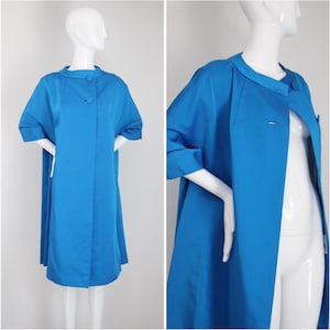 Puede incluir: Un abrigo vintage azul vibrante con cuello redondo y tres botones. El abrigo tiene mangas cortas y una silueta en forma de A. El abrigo se exhibe en un maniquí. Una pieza clásica de ropa de abrigo vintage.