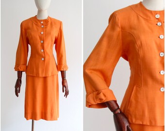 May include: A bright orange linen suit with a fitted jacket and a skirt. The jacket has a mandarin collar and long sleeves with button cuffs. The skirt is knee-length and has a straight silhouette.