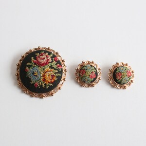 May include: A set of three vintage brooches featuring floral embroidery on a black background. The largest brooch is circular and has a gold-toned frame. The two smaller brooches are also circular and have a gold-toned frame.