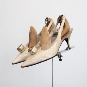 May include: A pair of vintage gold slingback heels with pointed toes. Each shoe features a decorative gold embellishment on the toe and a small heel. The shoes are displayed on a metal stand against a white background.