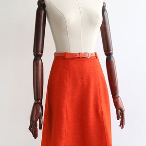 May include: A bright orange A-line skirt with a brown leather belt. The skirt is made of a textured fabric and has a simple, classic design.