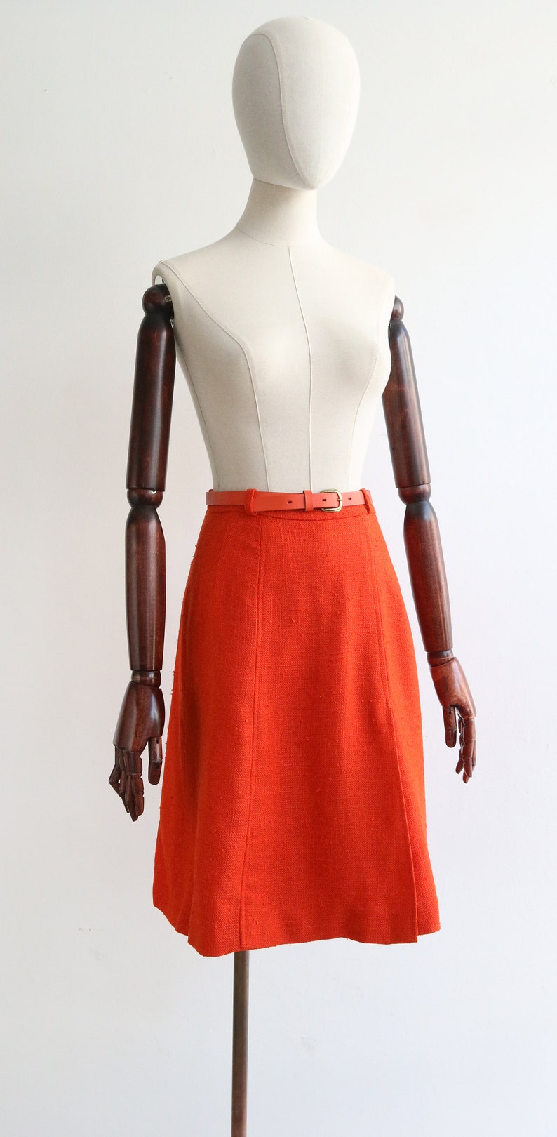 May include: A bright orange A-line skirt with a brown leather belt. The skirt is made of a textured fabric and has a simple, classic design.