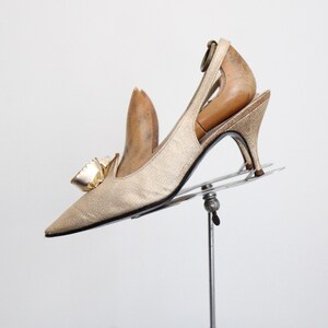 May include: A gold-colored, pointed-toe slingback heel with a bow detail. The vintage shoe has a slender heel and is displayed on a metal stand. The shoe is made of a shimmering fabric.