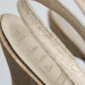 May include: Close-up of a pair of beige slingback heels. The interior sole displays the Christian Dior logo and the word "Souliers". The heels have a textured, shimmering appearance.