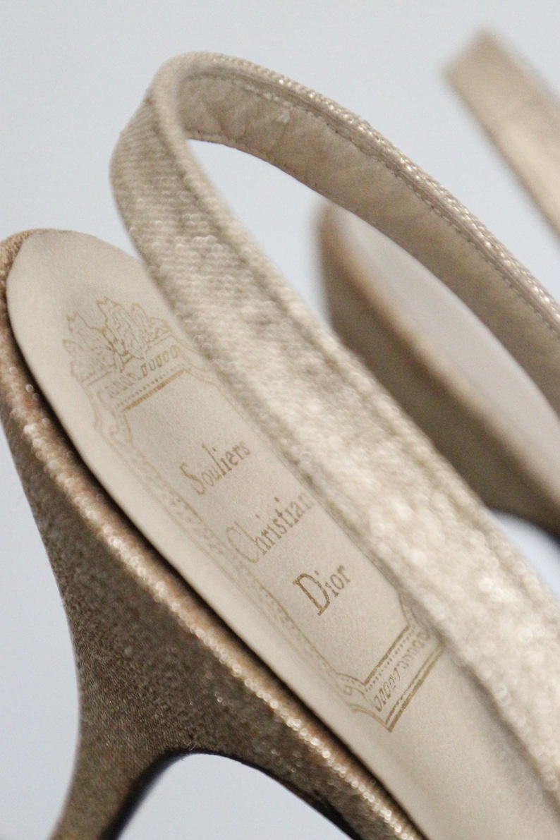 May include: Close-up of a pair of beige slingback heels. The interior sole displays the Christian Dior logo and the word "Souliers". The heels have a textured, shimmering appearance.