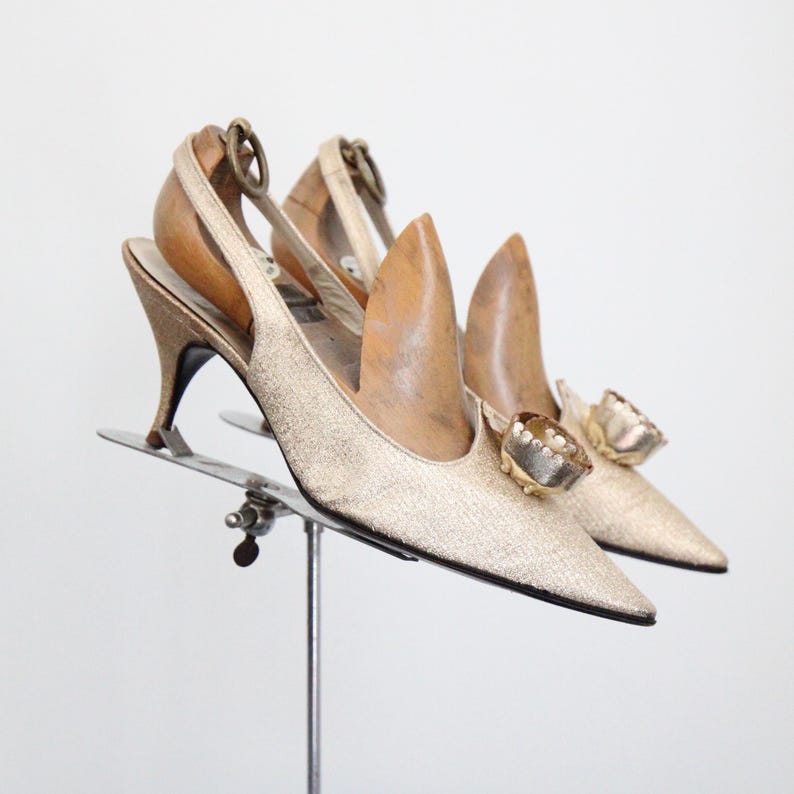 May include: A pair of gold slingback heels with pointed toes and small heels. The shoes have a metallic sheen and are adorned with decorative embellishments on the vamps. The heels are displayed on a metal stand.