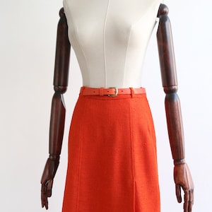 May include: A bright orange skirt with a brown leather belt. The skirt is knee-length and has a straight silhouette.