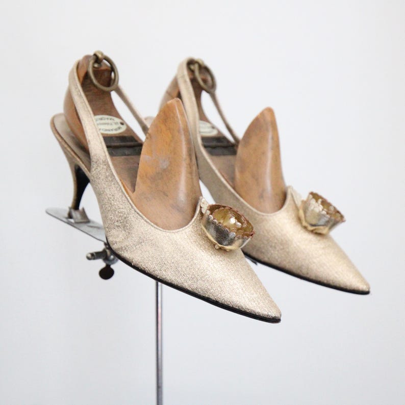 May include: A pair of vintage gold slingback heels with pointed toes. Each shoe features a decorative jewel on the vamp and a thin ankle strap. The heels are displayed on a metal stand against a white background.