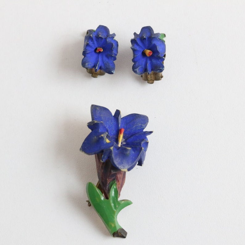 May include: A set of vintage floral jewelry, including a brooch and matching clip-on earrings. The brooch features a blue flower with a red and yellow center, a brown stem, and green leaves. The earrings mirror the flower design.