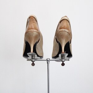 May include: A pair of gold-colored slingback heels with stiletto heels, displayed on a silver metal shoe stand. The shoes have a textured appearance and a wooden interior. The heels are approximately 3 inches tall.