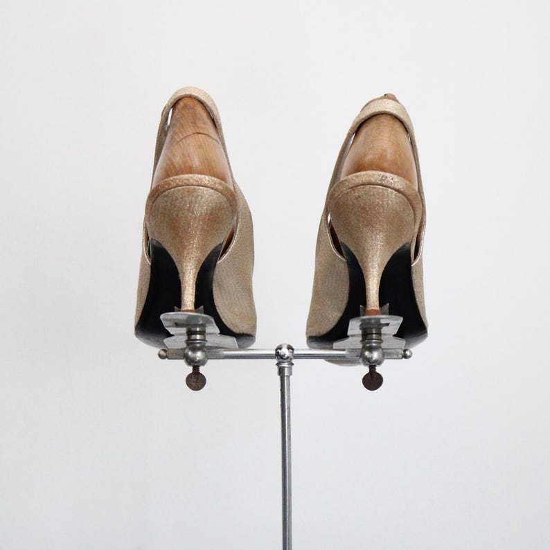 May include: A pair of gold-colored slingback heels with stiletto heels, displayed on a silver metal shoe stand. The shoes have a textured appearance and a wooden interior. The heels are approximately 3 inches tall.