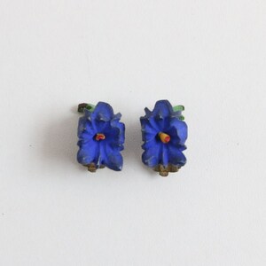 May include: A pair of vintage clip-on earrings shaped like blue flowers with yellow and red centers. The earrings have green leaves and gold-toned clips. The floral design is detailed and three-dimensional.