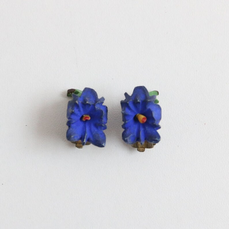 May include: A pair of vintage clip-on earrings shaped like blue flowers with yellow and red centers. The earrings have green leaves and gold-toned clips. The floral design is detailed and three-dimensional.