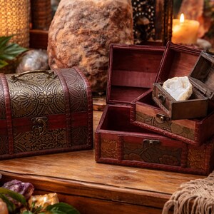 May include: A decorative wooden chest with ornate metal detailing and a handle sits next to a set of smaller open wooden boxes. The boxes are stained a dark red and are on a wooden surface. Candles and crystals surround the boxes.