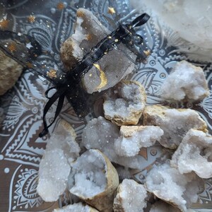 May include: A collection of quartz geodes and crystals, some with a white, crystalline interior. A black mesh bag with gold stars and crescent moon accents holds some of the crystals. The crystals are arranged on a decorative tray.