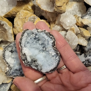 May include: A close-up of a light blue crystal geode with a dark gray outer layer. The geode is being held in a hand.