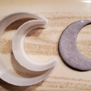 Op de afbeelding: Twee witte plastic maanvormige koekjesvormen en een zilveren glitter maanvormig koekje.