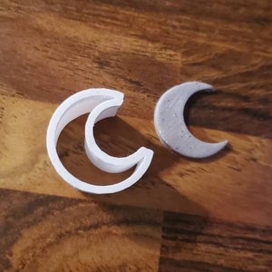 May include: Two white plastic crescent moon cookie cutters, one with a smaller crescent moon inside, and one gray plastic crescent moon cookie cutter.