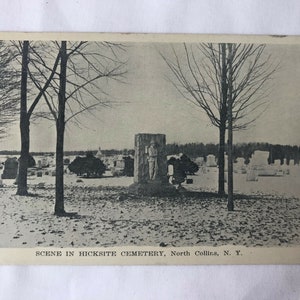 May include: Black and white photograph of a cemetery in the winter. The photo shows a snow-covered cemetery with a large stone monument in the center. The monument features a sculpture of a person standing on a pedestal. The text "SCENE IN HICKSITE CEMETERY, North Collins, N. Y." is printed at the bottom of the photo.