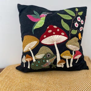 May include: A black pillow with embroidered mushrooms, a frog, a hummingbird, and leaves. The pillow is on a yellow chevron patterned throw blanket.