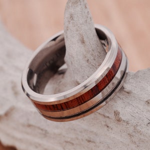 Può includere: Un anello d'argento con un intarsio in legno marrone rossiccio e una sottile fascia color oro. L'anello è appoggiato su un pezzo di legno trasportato dalle correnti. L'anello ha una finitura lucida e un design dettagliato.