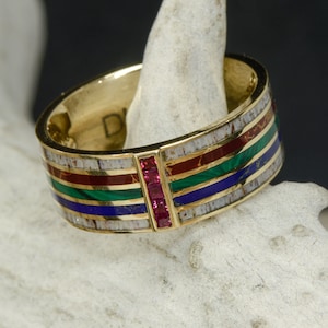 Pode incluir: Um anel de banda de ouro com uma incrustação multicolorida de pedras vermelhas, verdes, azuis e brancas. Uma fileira de pedras de rubi vermelho é engastada verticalmente no centro da banda.