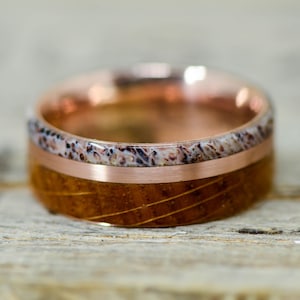 Può includere: Un anello di legno con una fascia in rame e un'intarsio bianco e marrone. L'anello è su una superficie di legno.