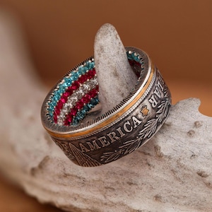 Pode incluir: Um anel de prata com uma banda dourada e um design de pedras preciosas vermelho, branco e azul. O anel é feito de uma moeda de meio dólar americano reutilizada e apresenta as palavras "LIBERTY" e "IN GOD WE TRUST".
