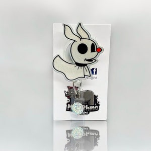 May include: A white glitter ghost character with a red nose and black eyes, attached to a retractable badge holder with a sparkly silver charm. The badge holder is attached to a white card with the text "Morphino Designs" in black.