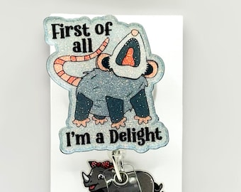 May include: A glittery acrylic badge reel featuring a gray opossum with its mouth open and the text First of all I'm a Delight. Below it is a smaller gray rhinoceros graphic with a red bow and the text Mrs. Rhino on a clear plastic strap.