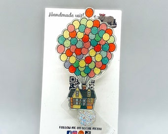 May include: A colorful, glittery resin charm featuring a house with balloons. The charm is attached to a white card with the text "Handmade with love" and "Follow me on social media @mrsrhinodesigns".