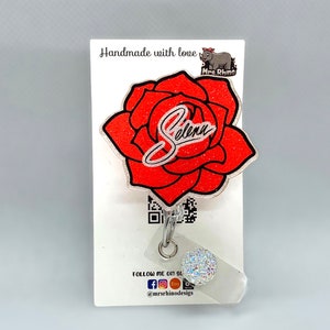 May include: A red glitter rose with the name "Selena" written in white glitter. The rose is attached to a silver key chain with a clear plastic charm that has a rainbow glitter design. The background is a white card with the text "Handmade with love Mrs. Rhino" and "Follow me on social @mrsrhinodesigns".
