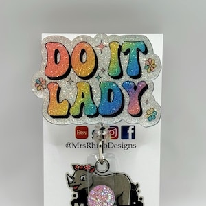 May include: A badge reel with the text "DO IT LADY" in rainbow glitter letters. Below is a gray rhino with a pink bow and a pink glitter ball. The Etsy shop name "MrsRhinoDesigns" is also visible.