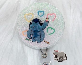 May include: A white glitter badge reel with a blue Stitch character drawing holding a crayon and surrounded by colorful hearts. The text "Mrs. Rhino Designs" is on the badge reel.