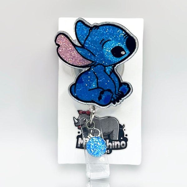 Può includere: Un portabadge blu glitterato raffigurante un personaggio con grandi orecchie e un dettaglio rosa all'interno. Il portabadge è fissato su un cartoncino bianco con un logo di un rinoceronte grigio e la scritta "Mishino Designs". Una piccola perla blu glitterata pende sotto.
