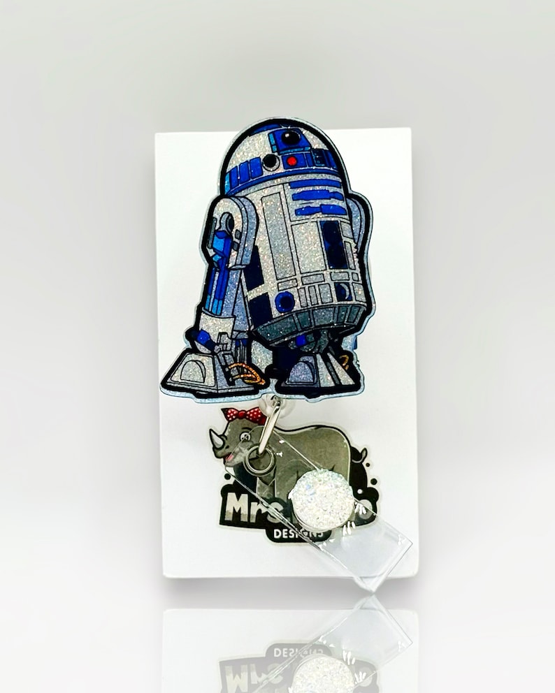 Op de afbeelding: Een badgehouder met een glitterende R2-D2 droid in blauw, zilver en wit. Onder de droid hangt een kleine grijze neushoornbedel met een rode strik met stippen. De badgehouder is bevestigd aan een witte displaykaart met de tekst Mrs. Designs.