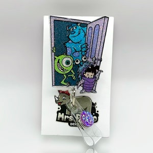 May include: A purple glitter badge holder with a clear strap and a purple glitter charm. The badge holder features a cartoon image of the characters from the movie Monsters Inc. The text "Mrs. Design" is visible on the badge holder.