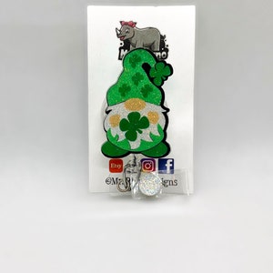 May include: A green glitter gnome with a shamrock on its hat and a white glitter beard. The gnome is attached to a retractable badge reel with a clear plastic cover and a silver metal ring. The reel is on a white card with the text "Etsy @Mr. Rhino Designs" and social media icons.