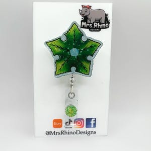 May include: A green and blue star-shaped badge reel with a glittery design. The badge reel has a clear plastic retractable cord and a green glitter accent. The top of the badge reel has a cartoon rhino logo and the text "Mrs. Rhino Designs".