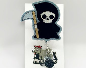 Puede incluir: Un dibujo animado en blanco y negro de la Muerte con una guadaña, sosteniendo un carrete de insignia de brillo negro y plateado con un encanto de brillo negro. El carrete de insignia está unido a una tarjeta blanca con el texto "Mrs. Designs".