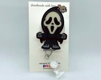 Puede incluir: Un portatarjetas con un personaje de dibujos animados con una máscara de fantasma blanca y una túnica negra con capucha. El personaje tiene la boca muy abierta y ojos negros. Debajo hay un anillo de metal plateado y una correa de plástico.