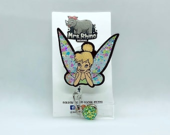 May include: A badge reel featuring a cartoon fairy with blonde hair in a bun and iridescent glitter wings. The character rests their chin on their hands. Below the fairy is a small, round, green glitter charm. The display card shows Mrs. Rhino Designs.