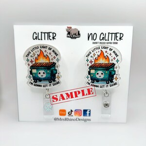 May include: Two badge reels featuring a dumpster fire illustration with the text "THIS LITTLE LIGHT OF MINE" and "I'M GONNA LET IT SHINE". One version has a glitter finish, while the other is plain. Both include a clear plastic clip and a metal button.