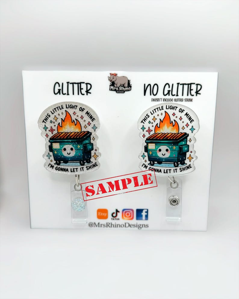 May include: Two badge reels featuring a dumpster fire illustration with the text "THIS LITTLE LIGHT OF MINE" and "I'M GONNA LET IT SHINE". One version has a glitter finish, while the other is plain. Both include a clear plastic clip and a metal button.