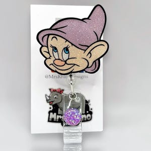 May include: A purple glitter badge reel with a cartoon character of a dwarf with a purple hat. The reel has a purple glitter charm with a clear background. The text "@MrsRhuDesigns" is printed on the card behind the reel.