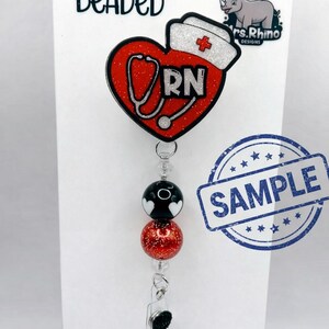 May include: A beaded badge reel featuring a red glitter heart with a white nurse cap, a stethoscope graphic, and the letters RN. The reel includes black and red glitter beads on a vertical cord. The display card reads BEADED, Mrs. Rhino DESIGNS, SAMPLE, and BEADED COLORS MAY VARY, BUT WILL MATCH THE DESIGN.